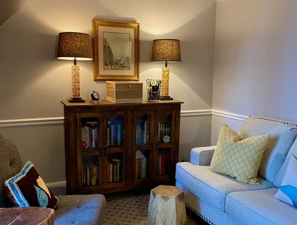 Living room corner with dark wood cabinet containing books, framed artwork, two lamps with brown shades, blue upholstered seating, and decorative pillows.